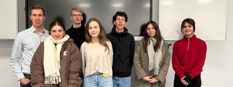 Sieben junge Erwachsene stehen in einem Klassenraum vor einem großen Bildschirm und weißen Tafeln.
