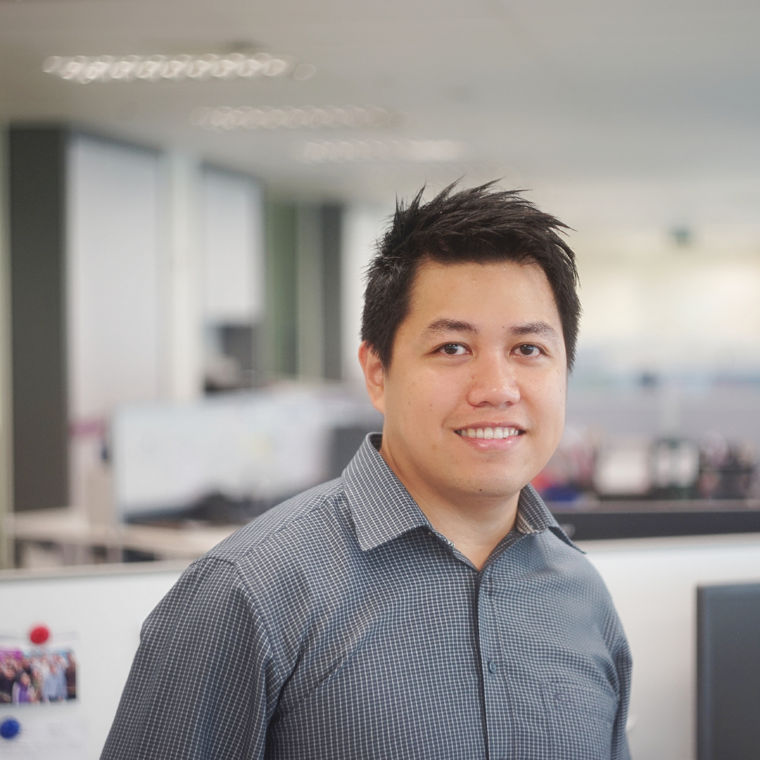 A person wearing a checkered shirt stands in an office environment with blurred workstations in the background.