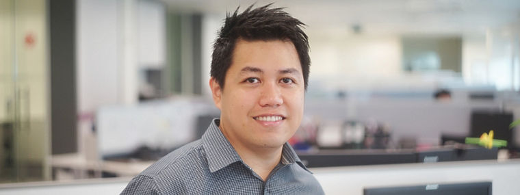 A person wearing a checkered shirt stands in an office environment with blurred workstations in the background.