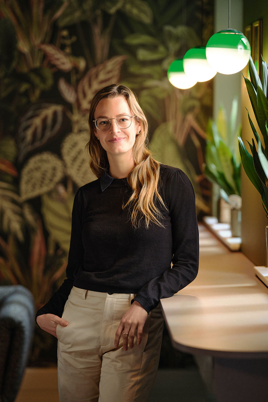 Myriam Rapior lehnt stehend an einem Sideboard und wird von oben rechts von kugelartigen Lampen angestrahlt