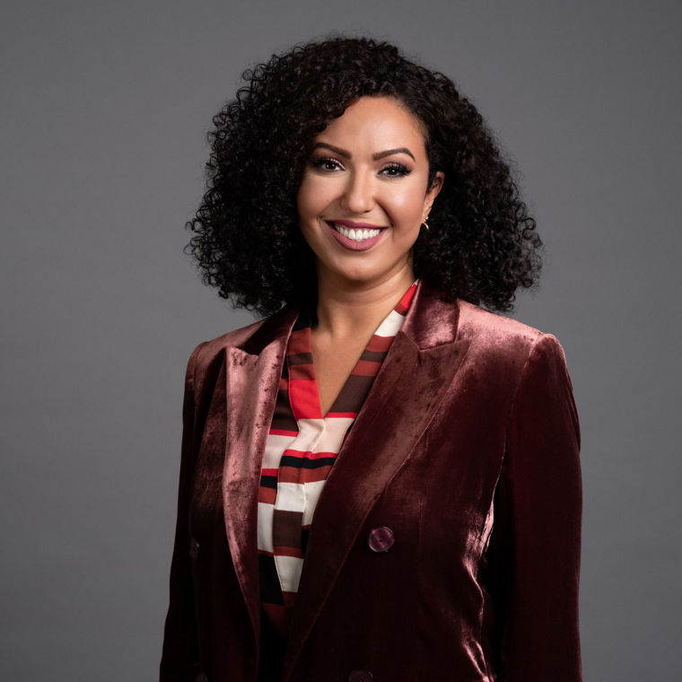 Woman with curly hair wearing a velvet maroon blazer and a patterned blouse, smiling against a gray background.