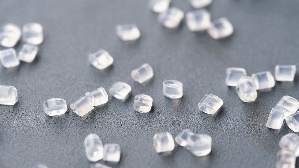 Small, clear cylindrical plastic granules scattered across a flat gray surface, representing raw polyamide material used to manufacture automotive components