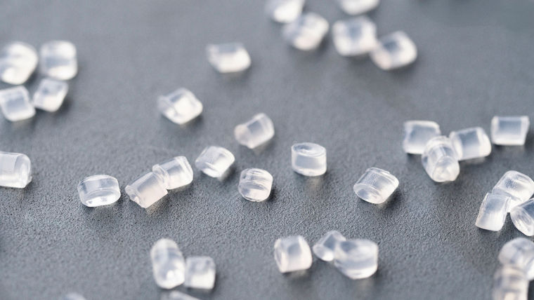Small, clear cylindrical plastic granules scattered across a flat gray surface, representing raw polyamide material used to manufacture automotive components