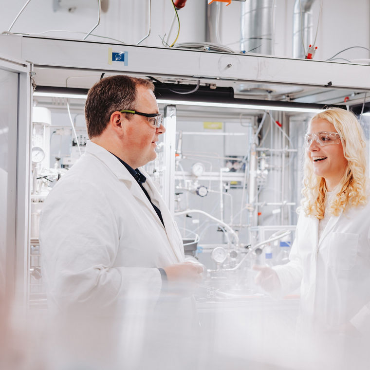 Andreas Vorholt und Lisa Steinwachs stehen in einem hellen Labor, beide tragen Laborkittel und Schutzbrillen und unterhalten sich.
