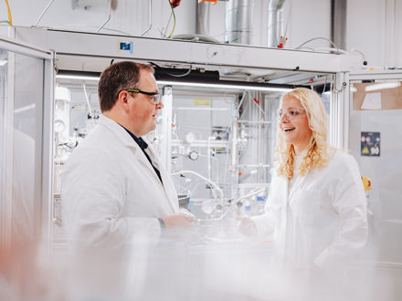 Andreas Vorholt and Lisa Steinwachs are standing in a bright laboratory, both wearing lab coats and safety goggles, and talking to each other.
