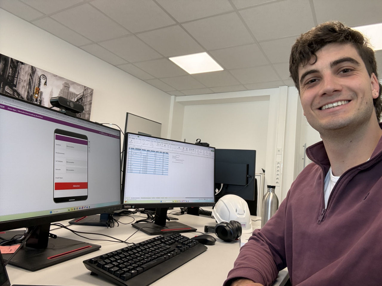 Man in purple sweater sitting at desk with dual monitors showing spreadsheet and app interface.