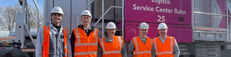 Five workers wearing orange safety vests and helmets stand in front of a purple train locomotive.