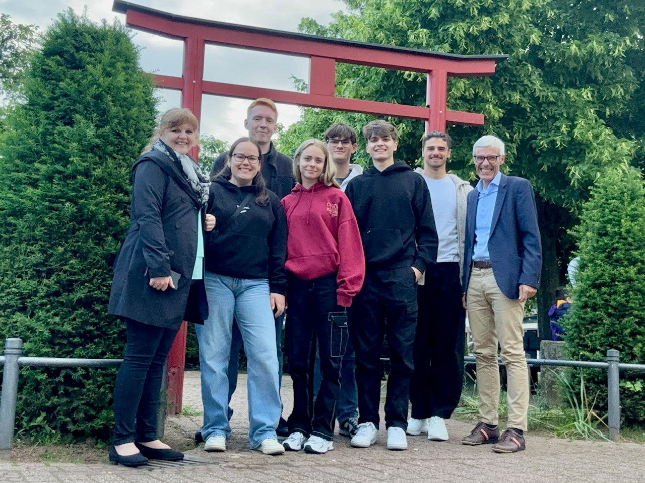 A group of seven people poses together in front of a red torii gate surrounded by greenery.