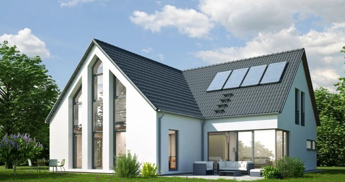 A modern two-story house with a dark gray sloped roof, white exterior walls, and large vertical windows, featuring rooftop solar panels and a patio with outdoor furniture, set in a lush green landscape under a partly cloudy sky.