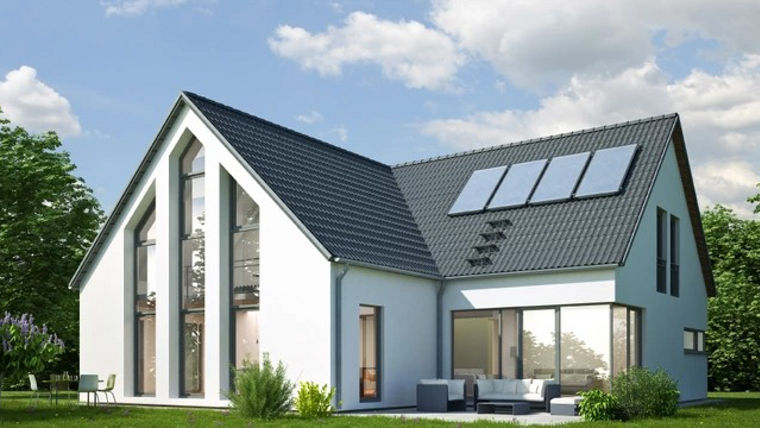 A modern two-story house with a dark gray sloped roof, white exterior walls, and large vertical windows, featuring rooftop solar panels and a patio with outdoor furniture, set in a lush green landscape under a partly cloudy sky.