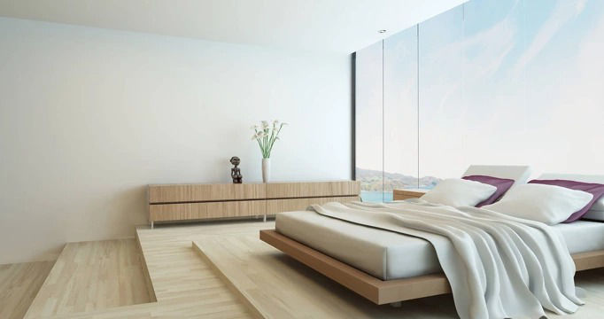 A modern, minimalist bedroom with light wooden flooring, a low platform bed dressed in white and purple, and a scenic view through a large window. 