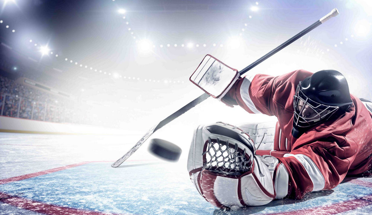 Hockey goalie in red gear diving to block a puck on an ice rink under bright lights.