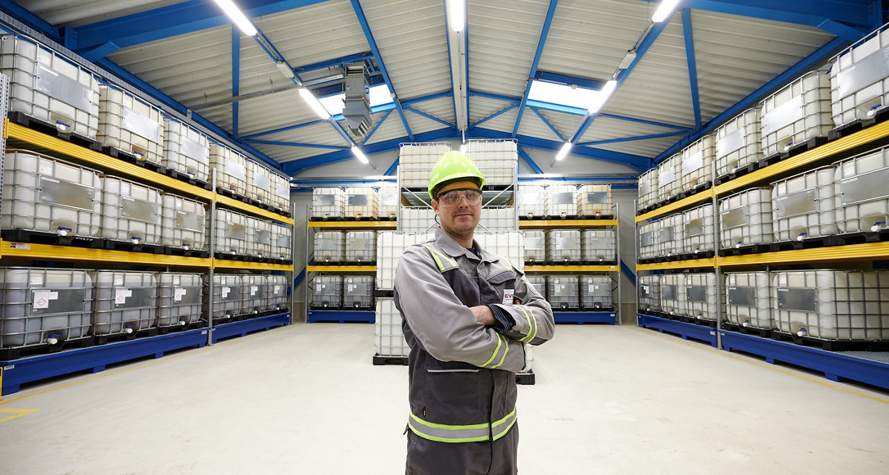 An employee in work clothes stands in the warehouse with his arms folded. Numerous stacked 1,000 liter canisters can be seen behind him.