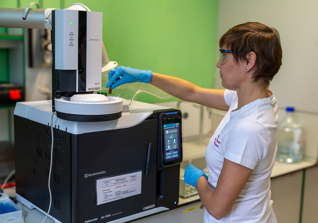 A laboratory technician inserting a sample into an analyzer. She is wearing white laboratory clothing, safety goggles and blue gloves. The sample vials are only a few centimeters high.