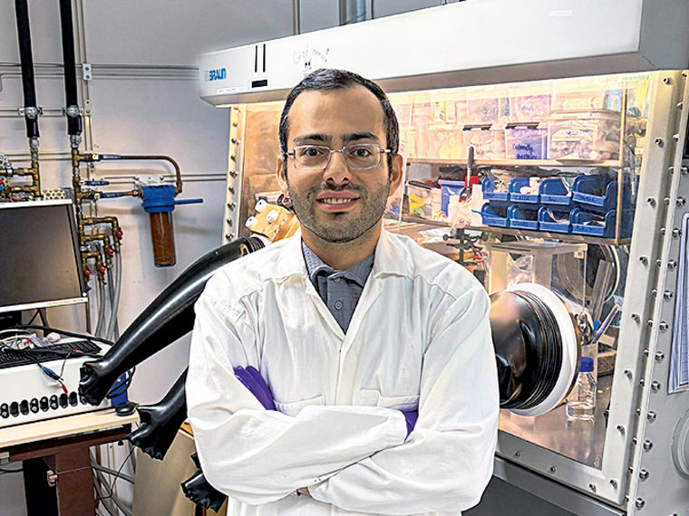 Saeed Mardi stands with arms crossed, wearing a lab coat, gloves, and safety goggles in a chemistry laboratory.