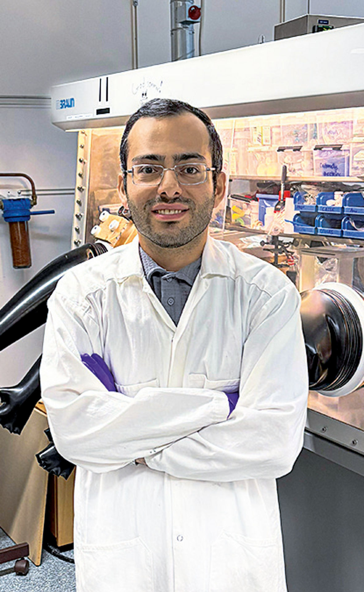 Saeed Mardi steht mit verschränkten Armen, Laborkittel, Handschuhen und Schutzbrille in einem Chemielabor.