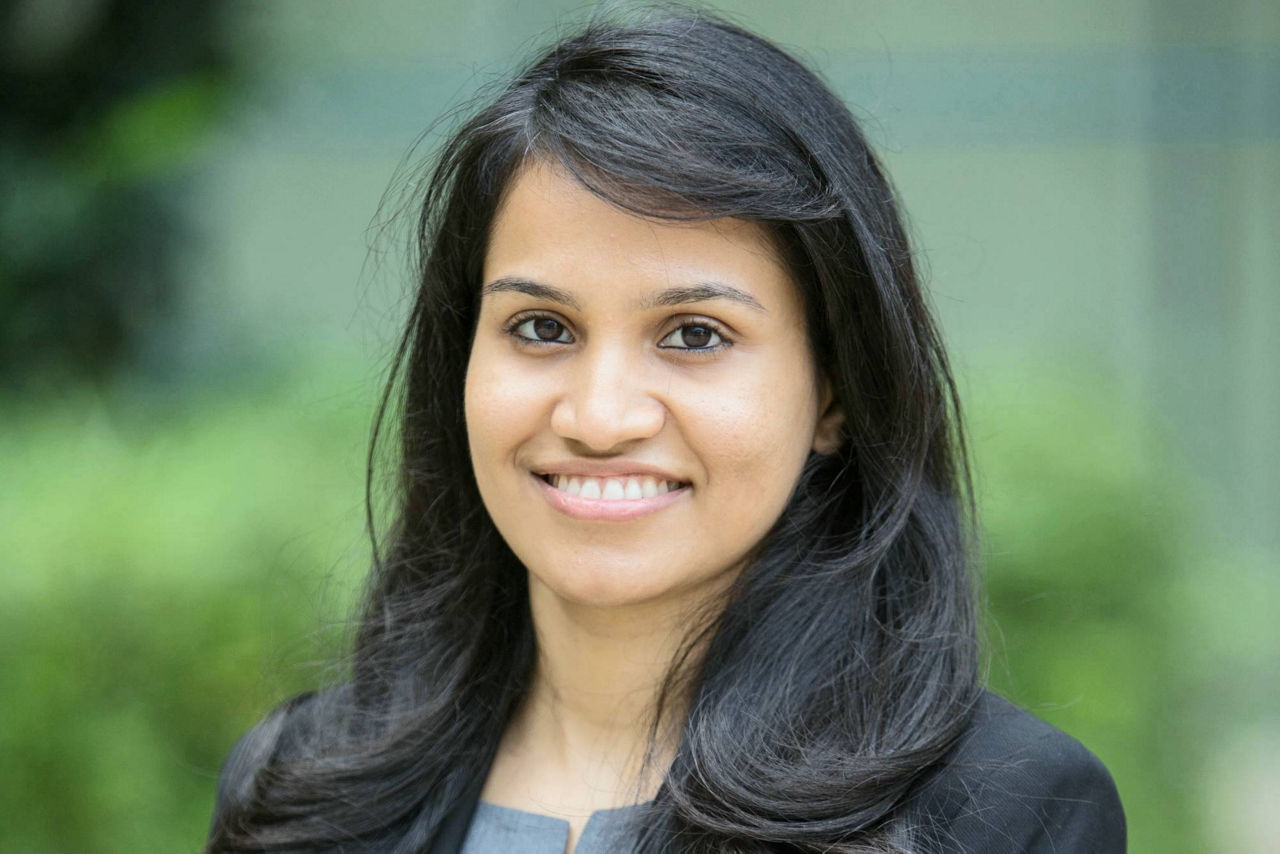 A woman with long black hair wearing a black blazer and grey top stands outdoors.