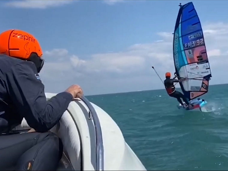 EIn Windsurfer, der von einem behelmten Mann in einem Motorboot verfolgt wird.