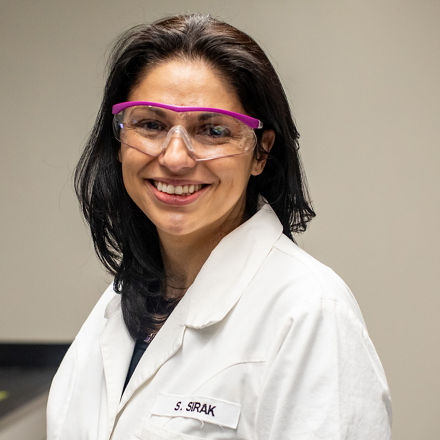 Sofia Sirak mit Laborbrille und Laborkittel lacht in die Kamera.