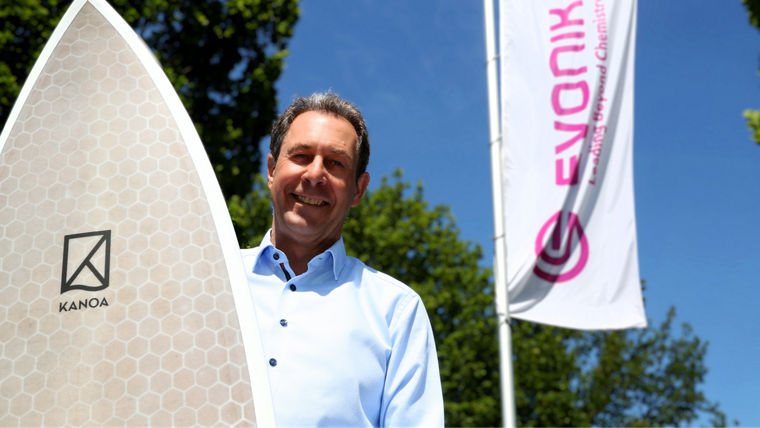 Stphan Sprenger with a KANOA surfboard and an Evonik flag behind him.