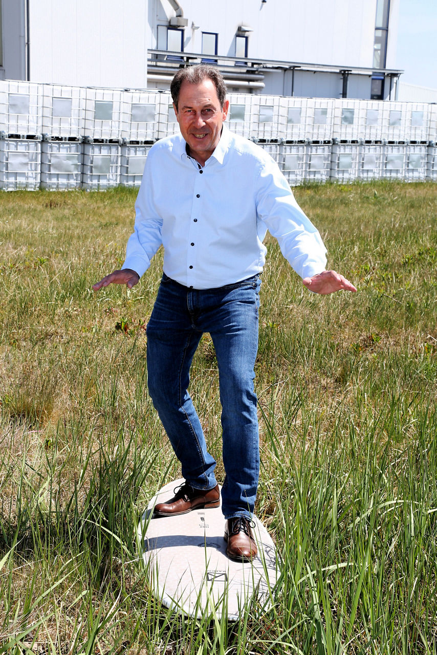 Stephan Sprenger steht auf einer Wiese auf einem Kanoa-Surfbrett. Im Hintergrund ist das Evonik-Werk in Geesthacht zu sehen.