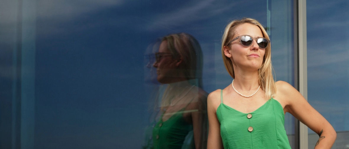 Woman in a green dress with wooden buttons stands confidently beside a reflective glass window.