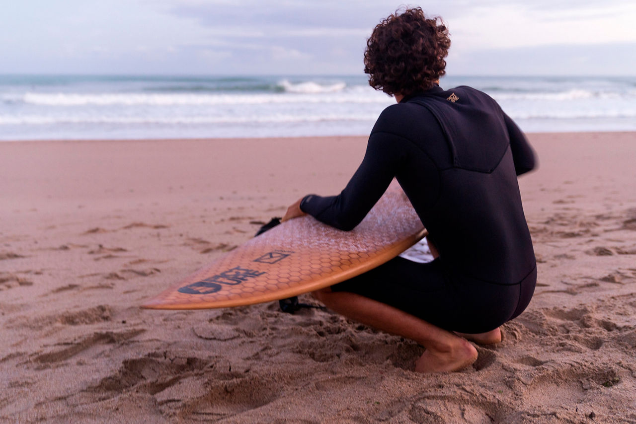 A surfer is crouching on the beach in a wetsuit, his surfboard resting on his thighs.