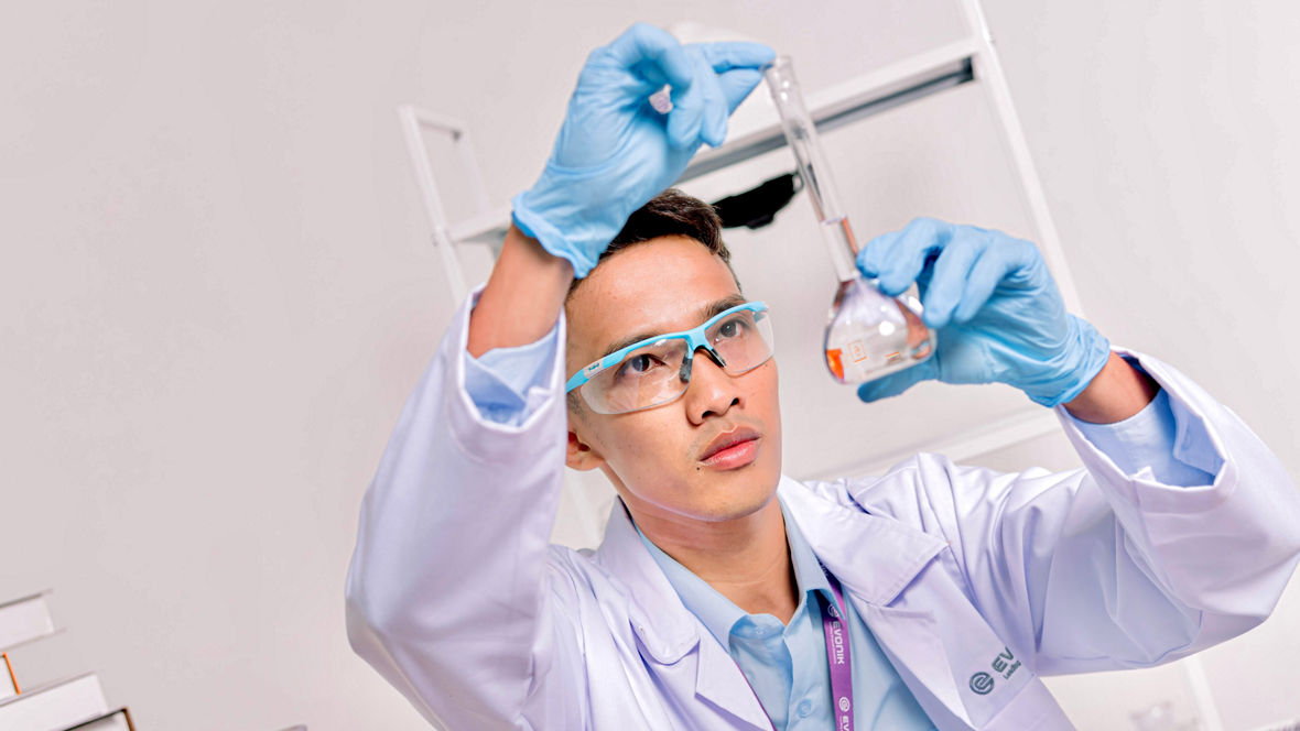 Scientist wearing blue gloves and a white lab coat holding a flask with orange liquid.