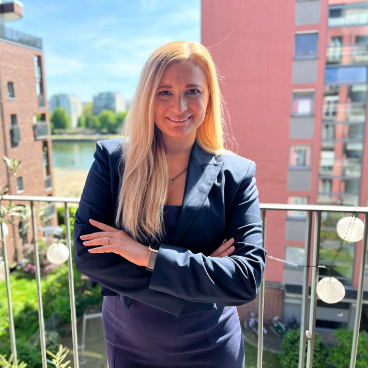 Frau mit langen blonden Haaren und blauem Blazer steht auf einem Balkon vor Wohngebäuden.