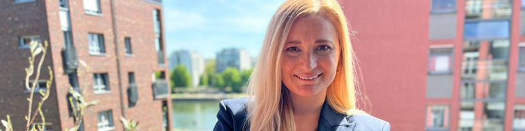 Person with long blonde hair wearing a dark blazer stands on a balcony with buildings in the background.