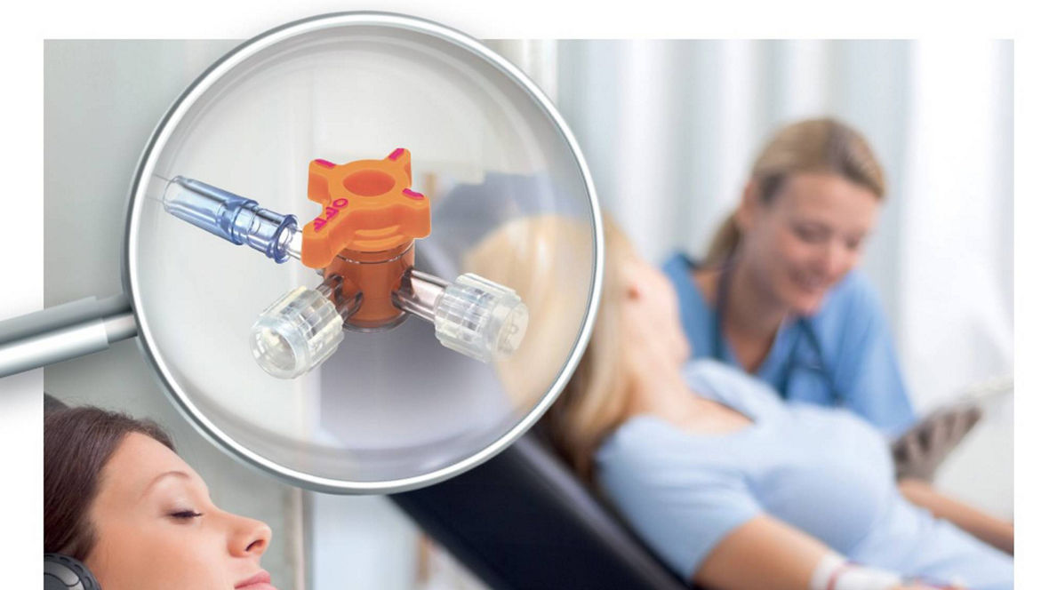 In a medical setting, a patient wearing headphones is reclining in a chair while receiving intravenous treatment. A magnifying glass highlights an orange medical device connected by tubes. In the background, a healthcare professional in scrubs is attending to another patient.