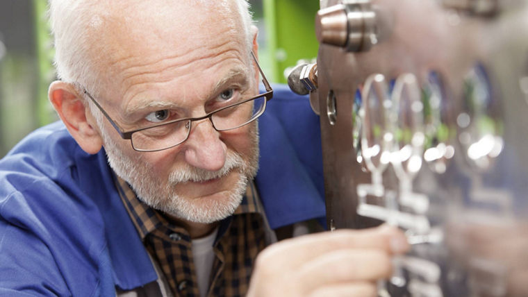 An elderly technician wearing a blue shirt and plaid undershirt is focused on adjusting machinery in a workshop, with his hands interacting with the equipment's metal panel featuring knobs and gauges.