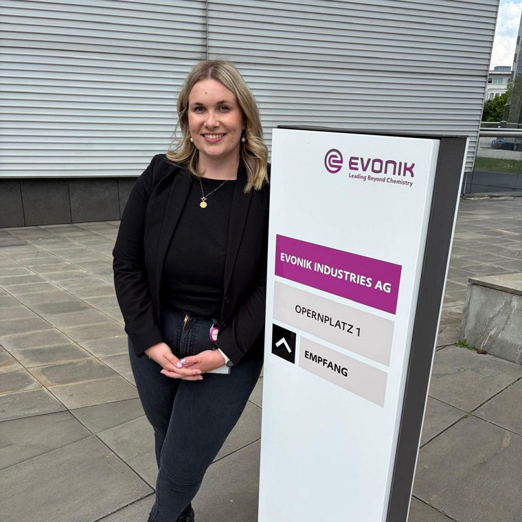 Woman in black jacket and jeans leaning against a tall white sign with purple and black text.