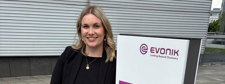 Woman in black jacket and jeans leaning against a tall white sign with purple and black text.
