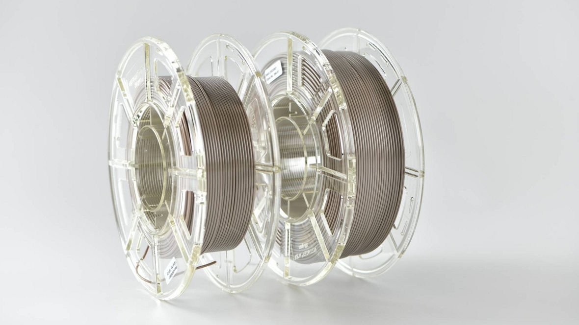 Two transparent spools of brown 3D printing filament on a white background. The filament is neatly wound around the central hubs, and the image is well-lit to highlight the material and structure of the spools.