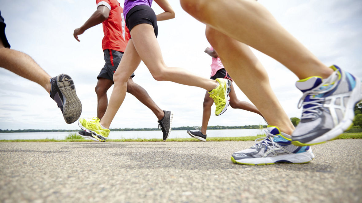Runners' legs and colorful shoes in motion on a paved path near a body of water.