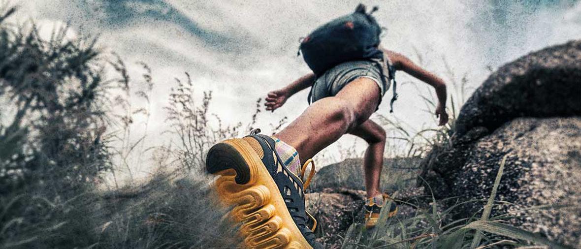A person hiking uphill wearing yellow-soled shoes and a backpack on rocky terrain.