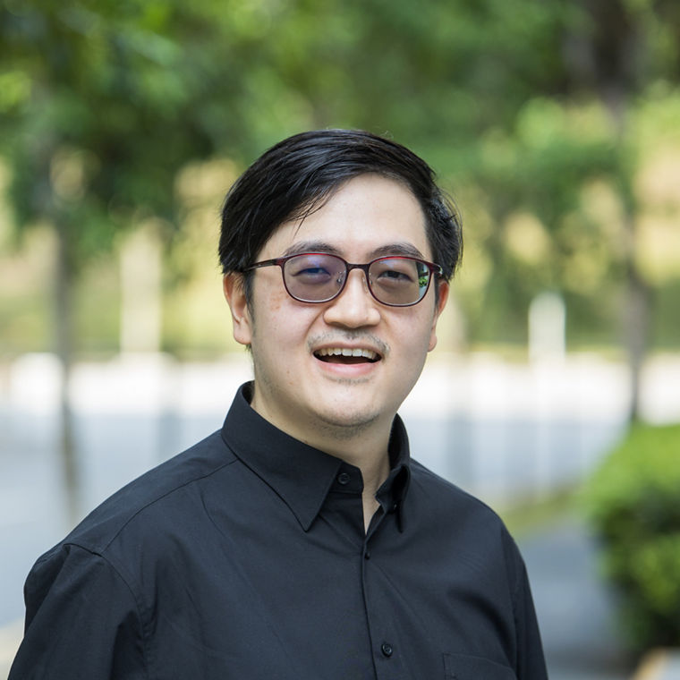Man wearing a black button-up shirt standing outdoors with blurred trees and road in the background.