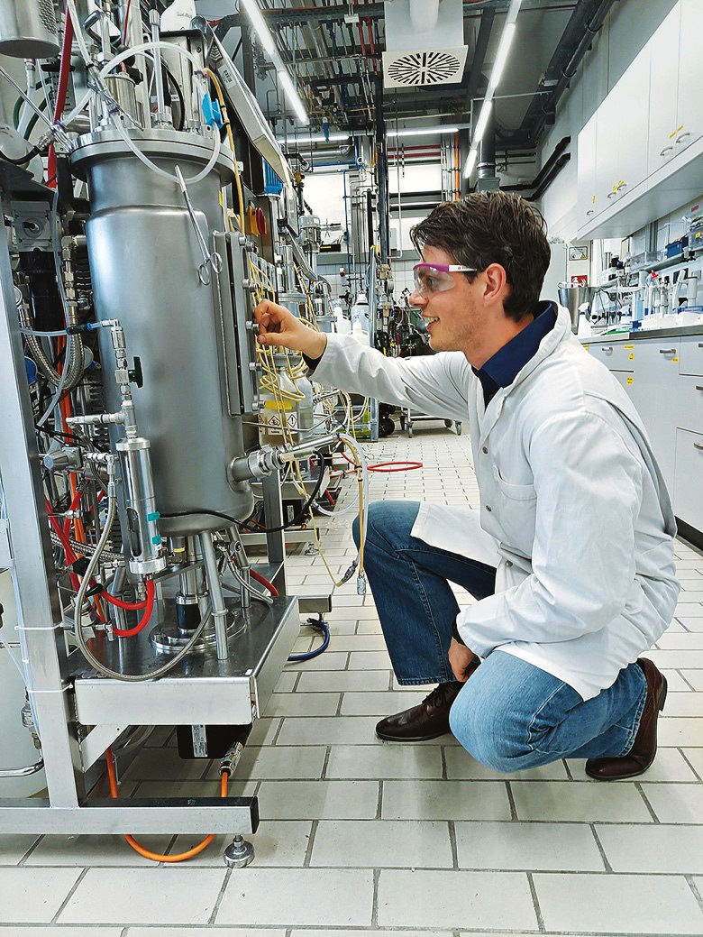 Xavier Hakkaart kneels in a lab coat and safety glasses in front of a fermenter about one meter high.