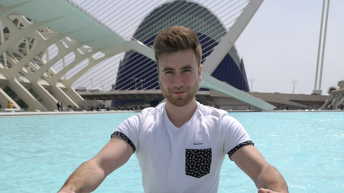 A person rowing a small boat in a turquoise body of water with modern architecture in the background.