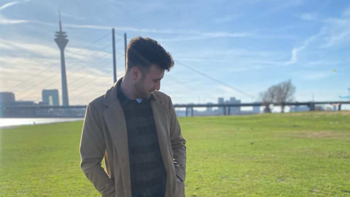 A person in a beige coat stands on green grass next to a small dog, with a city skyline in the background.