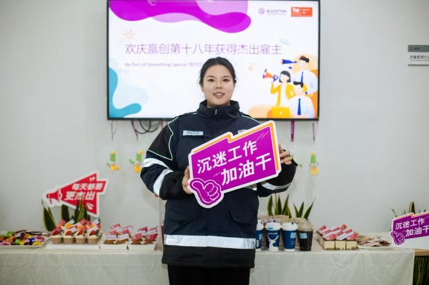 Eine Person in Arbeitskleidung hält ein Schild mit der Aufschrift „Arbeit vertiefen, Öl hinzufügen“ während einer Veranstaltung.