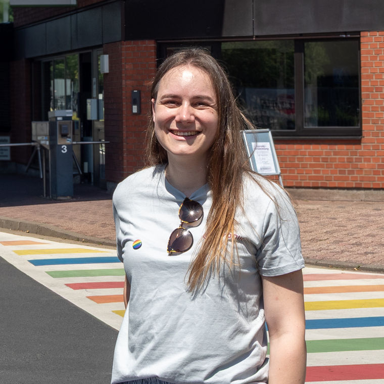 A smiling woman stands near a colorful striped crosswalk in front of a brick building.