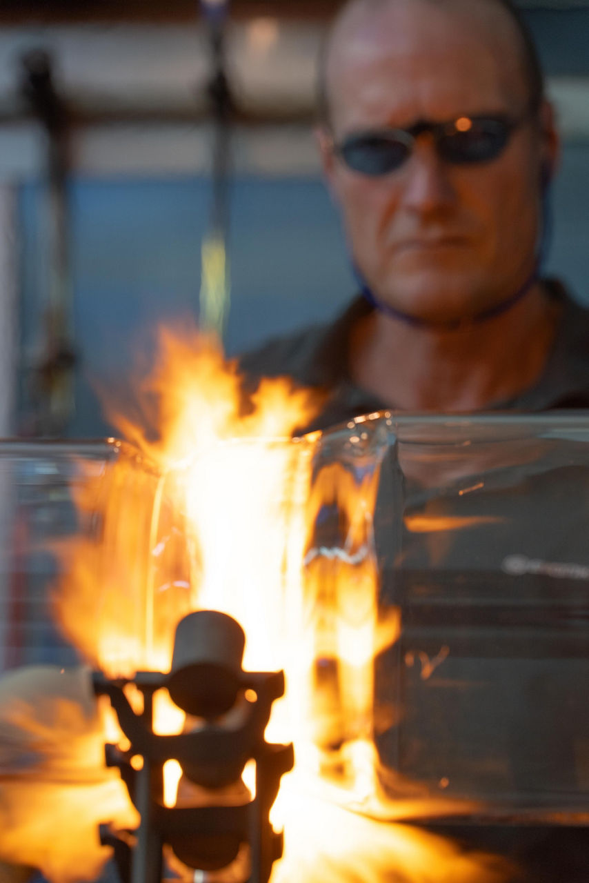 Udo Musholt beobachtet zwei Glaskörper, die sich in einer etwa 30 Zentimeter hohen lodernen Flamme treffen.