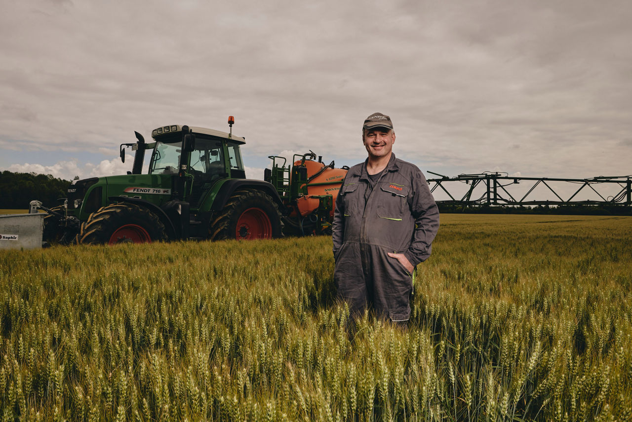 Ein Landarbeiter steht vor einem Traktor mit Sprüharmen auf einem Getreidefeld.