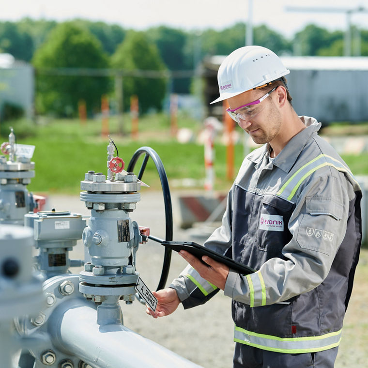 A person wearing a gray work uniform with yellow reflective strips and a white safety helmet featuring the Evonik logo is working on an outdoor industrial facility. The person is holding a tablet in one hand and operating a valve or device on a pipeline with multiple fittings using the other hand. In the background, green trees, blur effects, and construction fencing are visible.