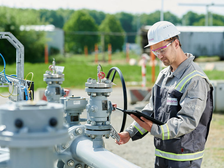Eine Person in einer grauen Arbeitsuniform mit gelben Reflektorstreifen und einem weißen Schutzhelm, der das Unternehmenslogo „Evonik“ trägt, arbeitet an einer industriellen Anlage im Freien. Die Person hält ein Tablet in einer Hand und bedient mit der anderen Hand ein Ventil oder Gerät an einer Rohrleitung mit mehreren Armaturen. Im Hintergrund sind grüne Bäume, Unschärfeeffekte und Bauzäune sichtbar.