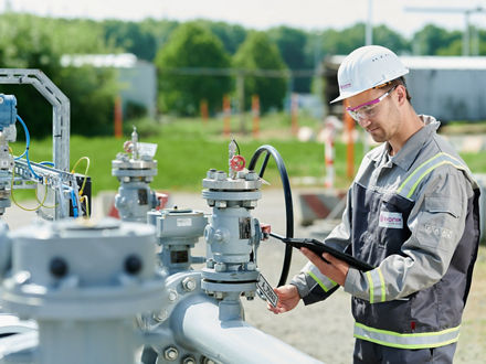 A person wearing a gray work uniform with yellow reflective strips and a white safety helmet featuring the Evonik logo is working on an outdoor industrial facility. The person is holding a tablet in one hand and operating a valve or device on a pipeline with multiple fittings using the other hand. In the background, green trees, blur effects, and construction fencing are visible.