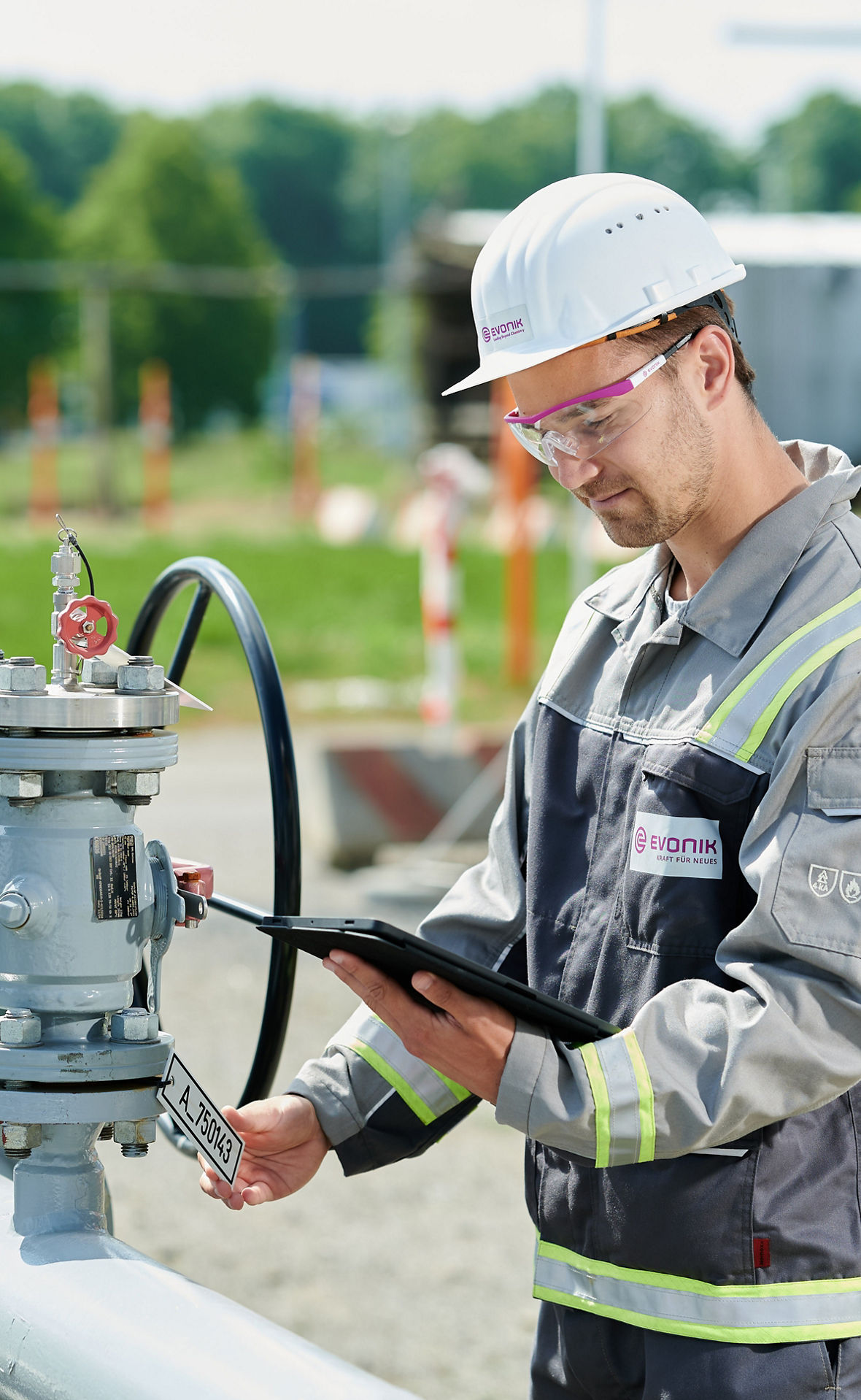 Eine Person in einer grauen Arbeitsuniform mit gelben Reflektorstreifen und einem weißen Schutzhelm, der das Unternehmenslogo „Evonik“ trägt, arbeitet an einer industriellen Anlage im Freien. Die Person hält ein Tablet in einer Hand und bedient mit der anderen Hand ein Ventil oder Gerät an einer Rohrleitung mit mehreren Armaturen. Im Hintergrund sind grüne Bäume, Unschärfeeffekte und Bauzäune sichtbar.