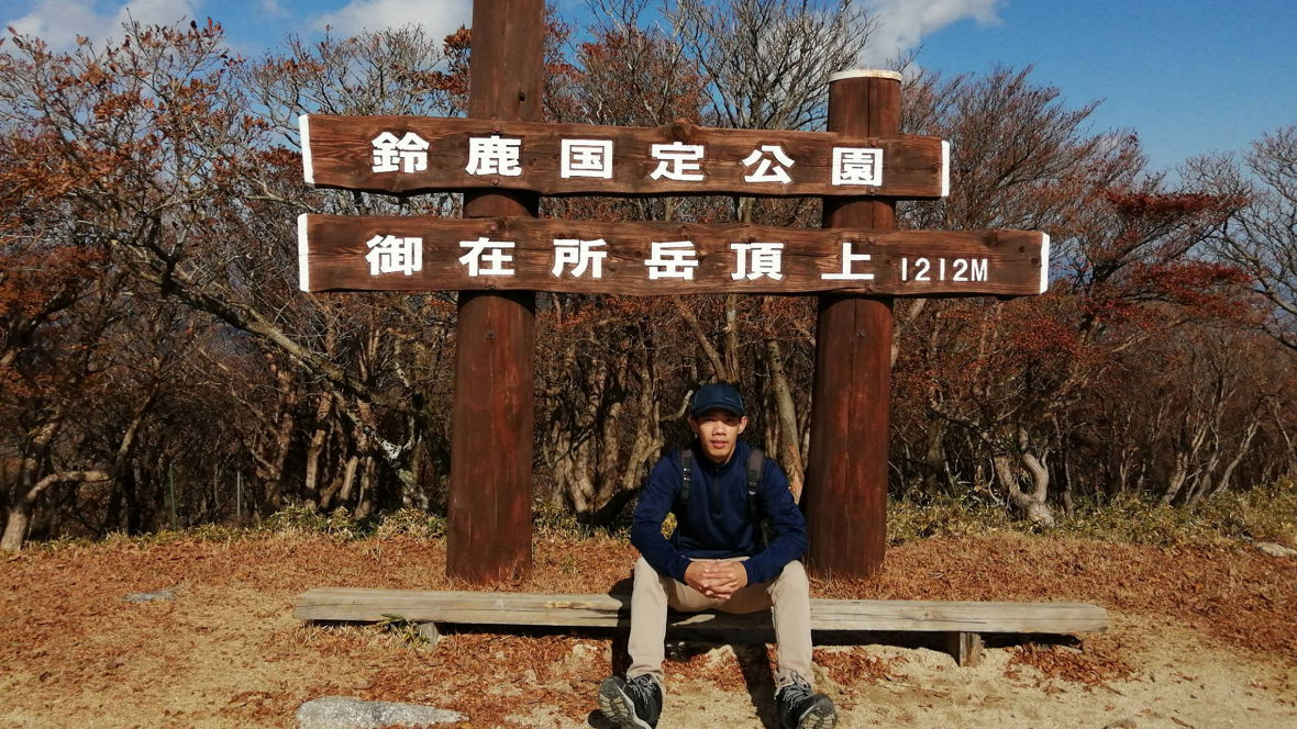 A person sitting on a bench in front of a sign indicating the summit of a mountain in Suzuka National Park, Japan.